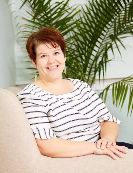 Jess, a white woman with short hair sits comfortably in front of a green plant. She has dimples and smiles at the camera. She is wearing a white short-sleeve top with blue stripes and dangly silver earrings. 
