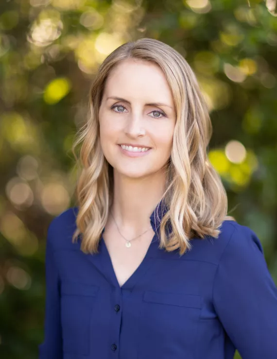 Professional headshot of blonde woman in blue shirt standing with trees