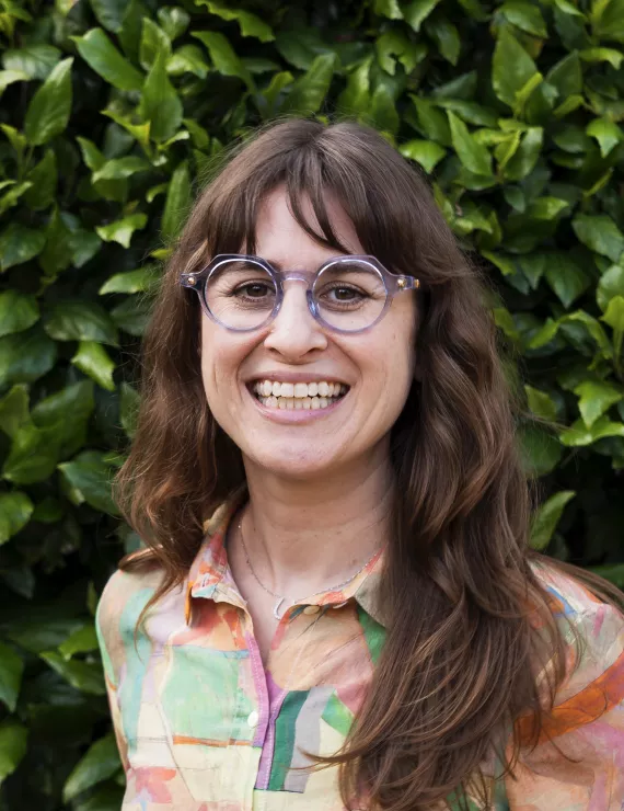Female Therapist in front of leafy green background smiling, brown hair, glasses, and colorful button down shirt.
