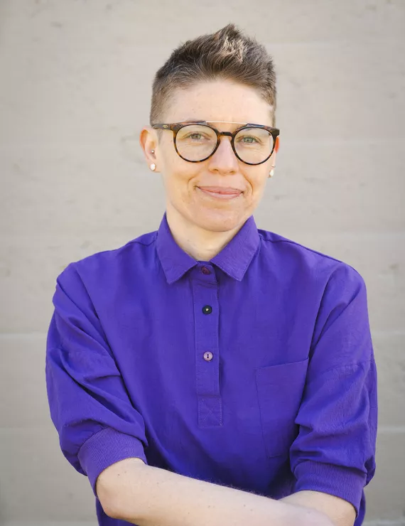 Ashley is a thin queer white woman with very short hair wearing a purple short sleeve-shirt and large-rimmed glasses has her arms crossed in front of her.