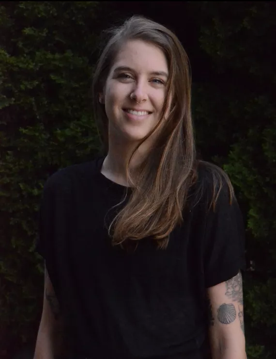 Kay is pictured center frame from head to torso. Kay has long hair worn down and to the side with a black short sleeve shirt. Kay is smiling, tattoos can be seen on their left arm. A wall of leaves is behind them. 