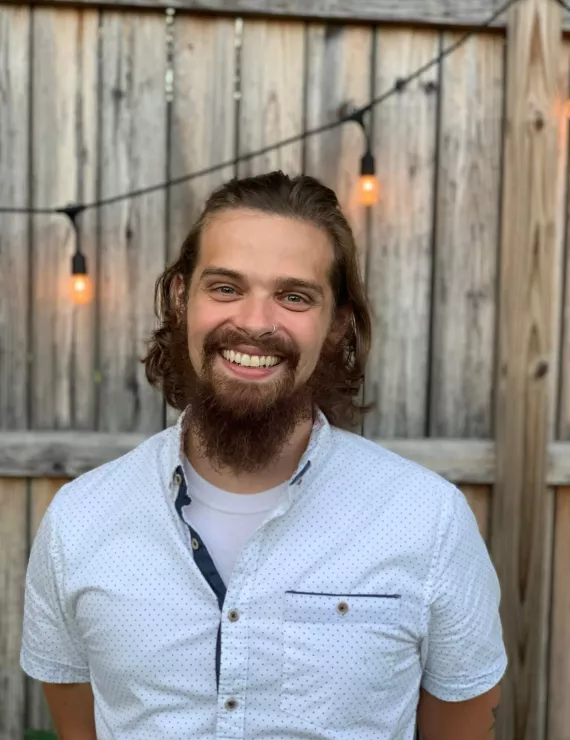 Image: Me, a medium-height male-presenting person. My long brown hair is worn up. I am wearing a white, blue spotted shirt with a collar. Behind me, there is a brown fence with string lights dangling from it. 