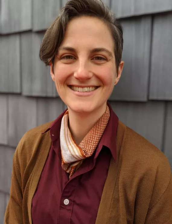 Photo of Erika - white, non-binary, brown short hair, looking at camera with gray shingles in background. Erika wearing maroon button up shirt and gold sweater with a polka dot handkerchief.