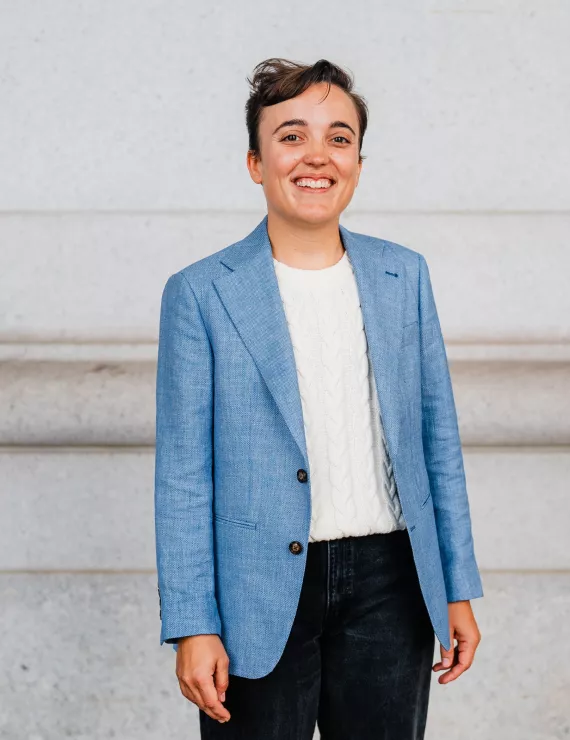 A smiling person standing in front of cream columns wearing a blue blazer and jeans. 