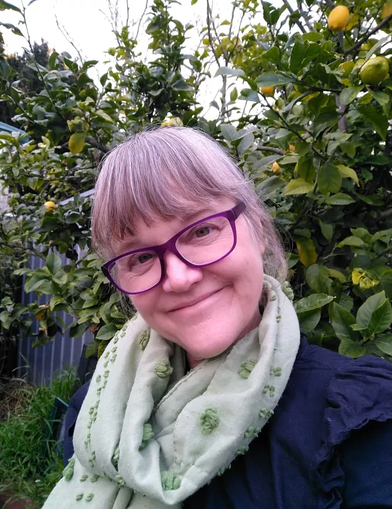 Picture of Edwina in a garden smiling, wearing glasses and a scarf.
