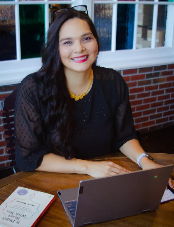 Achara sitting at a table smiling with a laptop in front of her