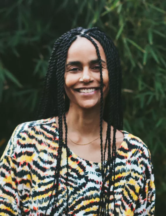 Smiling brown skinned woman with long braids wearing a tiger print shirt standing in front of a tree