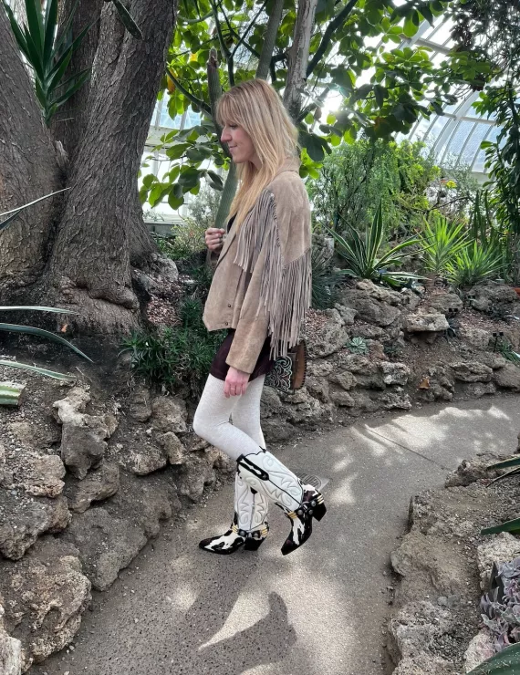 Blonde haired woman wearing a fringe jacket and cowboy boots surrounded by plants