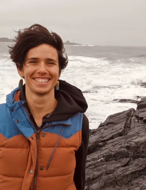 Ana stands in front of a stormy Maine sea and rock outcropping. She is white with olive skin and short brown hair and she is wearing a brown and blue vest on top of a black sweatshirt.