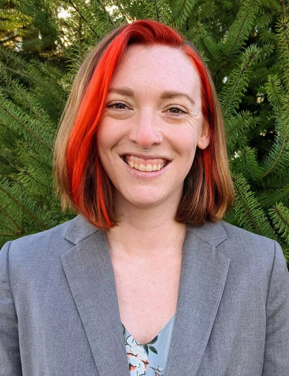 A smiling femme presenting white person with red hair wearing a grey blazer. 