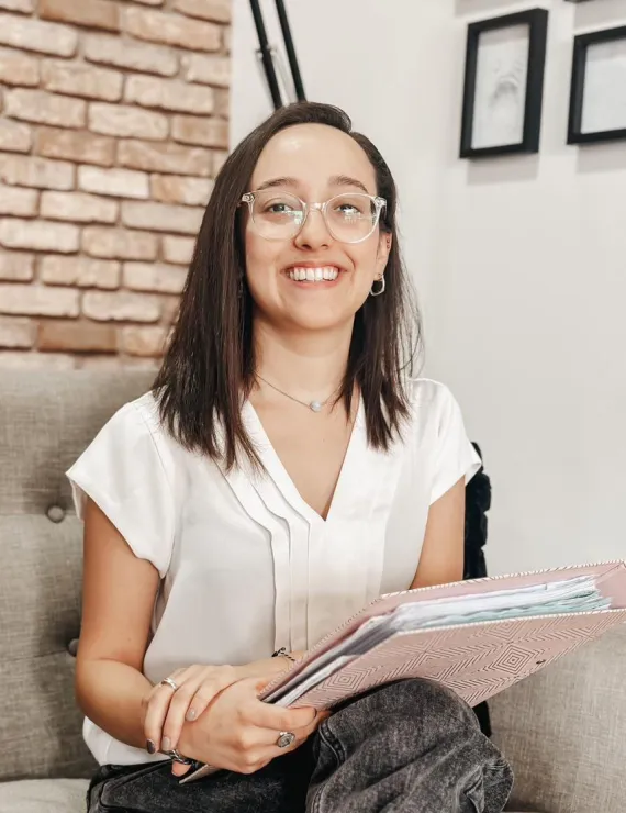 Una mujer joven está sentada en un sillón gris claro, sonriendo de forma cálida y amigable. Tiene el cabello lacio, castaño oscuro y a la altura de los hombros. Lleva gafas transparentes y redondeadas, y usa una blusa blanca con pliegues en el centro. También lleva un collar delicado con un dije pequeño y redondo. Sostiene una carpeta rosada con patrones geométricos y varias hojas dentro. Está en un ambiente acogedor: a su lado hay una pared de ladrillos y en el fondo se ven cuadros enmarcados.