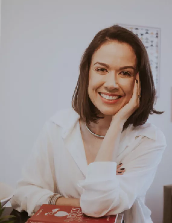 Mulher de 35 anos, de cabelos castanhos e curtos, com mão esquerda no rosto sorrindo. 