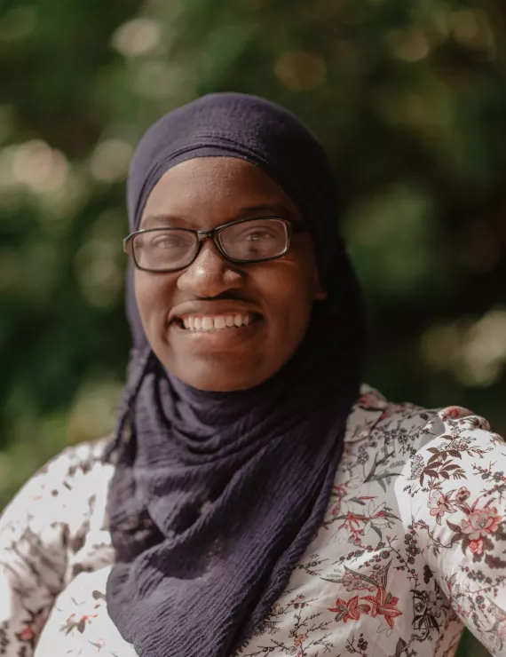 Fatimah is pictured from the chest up slightly angled toward the camera. She is wearing a blue headscarf, a floral print shirt, black glasses and a smile that shows her teeth.